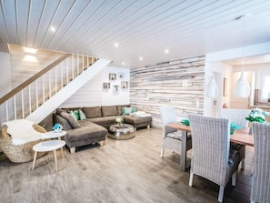 A modern living room with a wooden staircase, a sectional sofa, a dining area with wicker chairs, and a stone wall. - A gem of a Modern Retreat (Oberharz am Brocken)