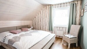 A bedroom with a sloped ceiling, a bed with a quilted cover, a chair, and a window with curtains. - A gem of a Modern Retreat (Oberharz am Brocken)