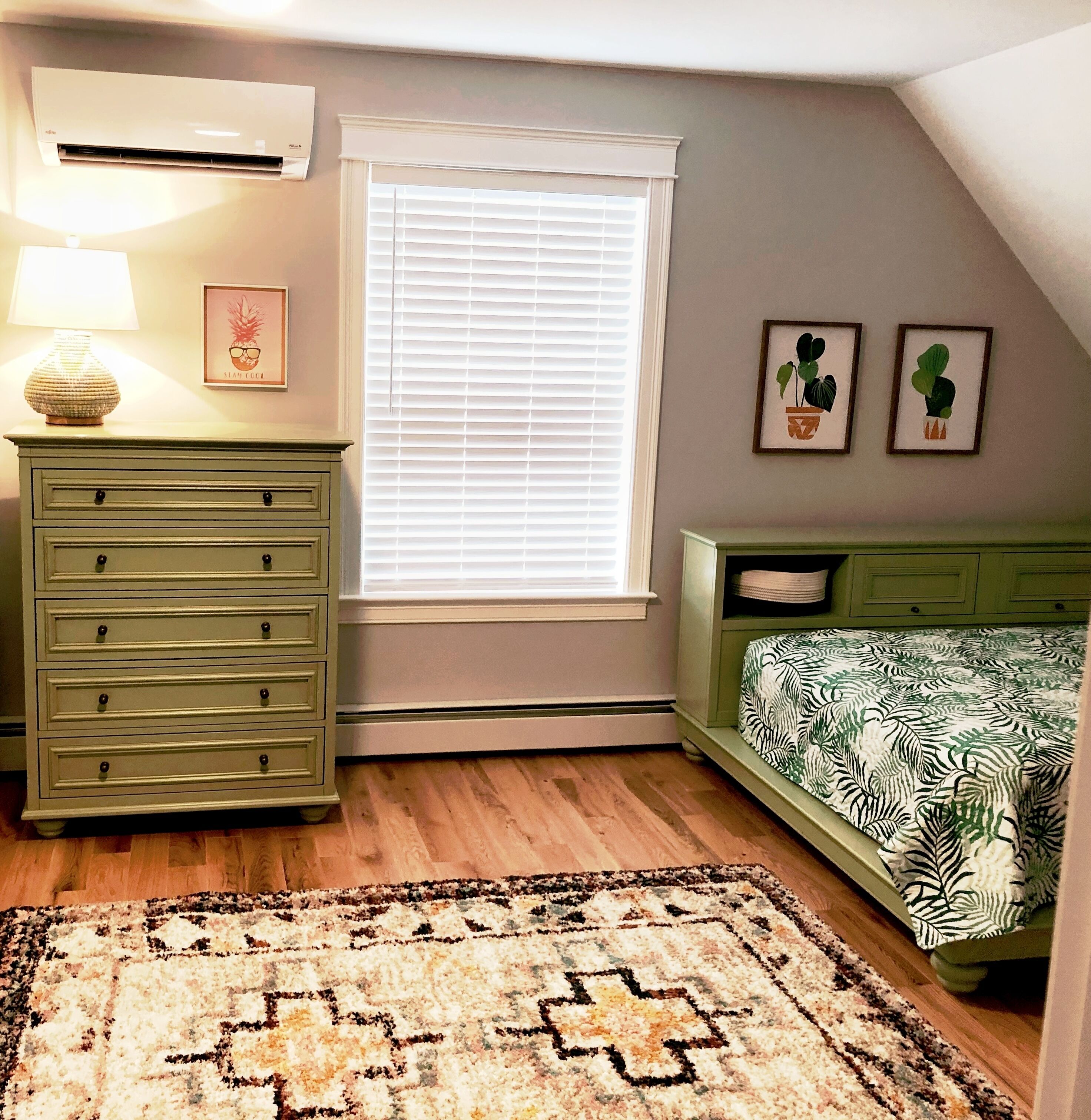 A bedroom with a green dresser, a bed with a patterned bedspread, a window with blinds, a wall-mounted air conditioner, and a rug.