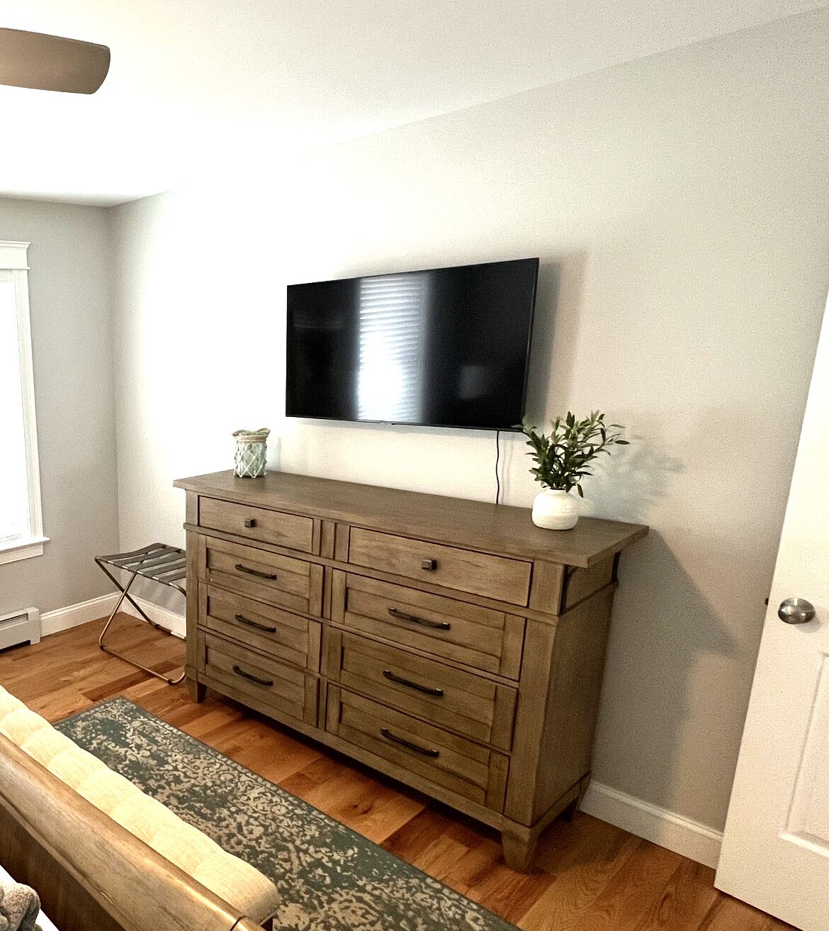 A room with a wooden dresser, a flat-screen TV mounted above it, a small potted plant, and a window with blinds.