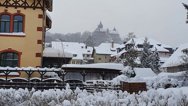 A traditional European building with a steep roof and a prominent tower, surrounded by snow-covered houses and a castle in the background. - Apartment 1 in an Art Nouveau Villa (Wernigerode)