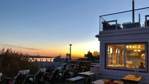 An outdoor seating area with tables and chairs, overlooking a marina with boats and a sunset in the background.