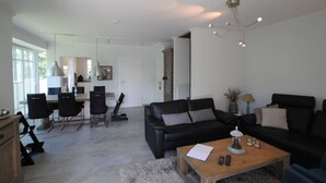 A modern living room with a black leather sofa, a wooden coffee table, and a dining area with a white table and black chairs. - Residence Island Pearl (Langeoog)