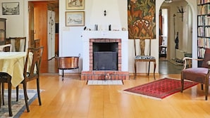 Sala de estar com lareira, piso de madeira, mesa de jantar, cadeiras, tapete vermelho e estante de livros.