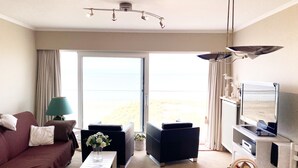 A hotel room with a sofa, a small table with a vase of flowers, a TV, and a view of the beach. - Apartment in Nieuwpoort Near the Beach (Nieuwpoort)