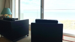 A room with a large sliding glass door leading to a balcony with a view of the sea. There is a dark-colored sofa and a lamp on a side table. - Apartment in Nieuwpoort Near the Beach (Nieuwpoort)