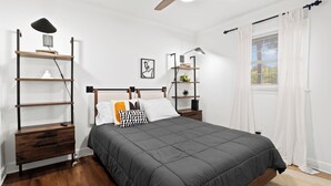 A bedroom with a bed, a ceiling fan, a nightstand, a desk with a lamp, and a window with curtains.