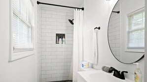 A modern bathroom with a white tiled shower, a toilet, a sink with a mirror, and a window with blinds.