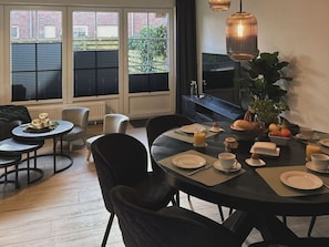 Un comedor con una mesa redonda lista para el desayuno, con platos, tazas y diversos alimentos. Hay sillas alrededor de la mesa, y la habitación tiene ventanas amplias con persianas.