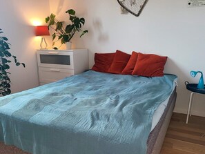 Een slaapkamer met een bed, nachtkastje, lamp, plant en een klein tafeltje met een blauw voorwerp.