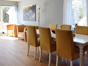 Een eethoek met een witte tafel en bijpassende stoelen, een schilderij aan de muur en uitzicht op bomen door het raam.