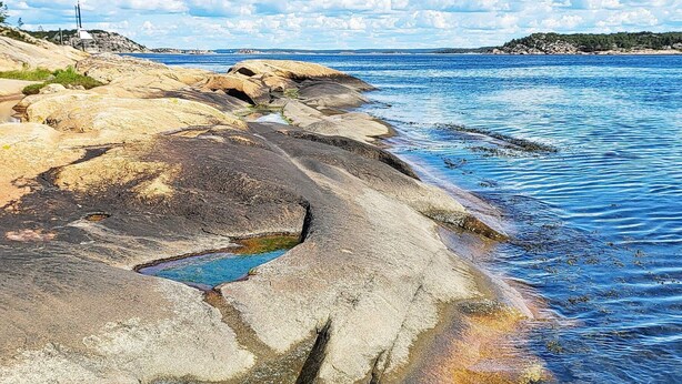 A rocky shoreline with clear blue water and a distant view of a lighthouse. - 7 Person Holiday Home in Vesteroy-by Traum (Hvaler)