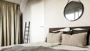 Een net opgemaakt bed met een gestreept deken, een ronde spiegel aan de muur en een ladder tegen de deur geleund.