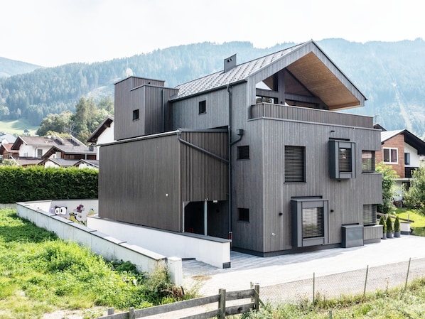 Une maison moderne à plusieurs étages, dotée d’un toit en pente, de grandes fenêtres et d’un espace extérieur visible, délimité par une clôture en bois.