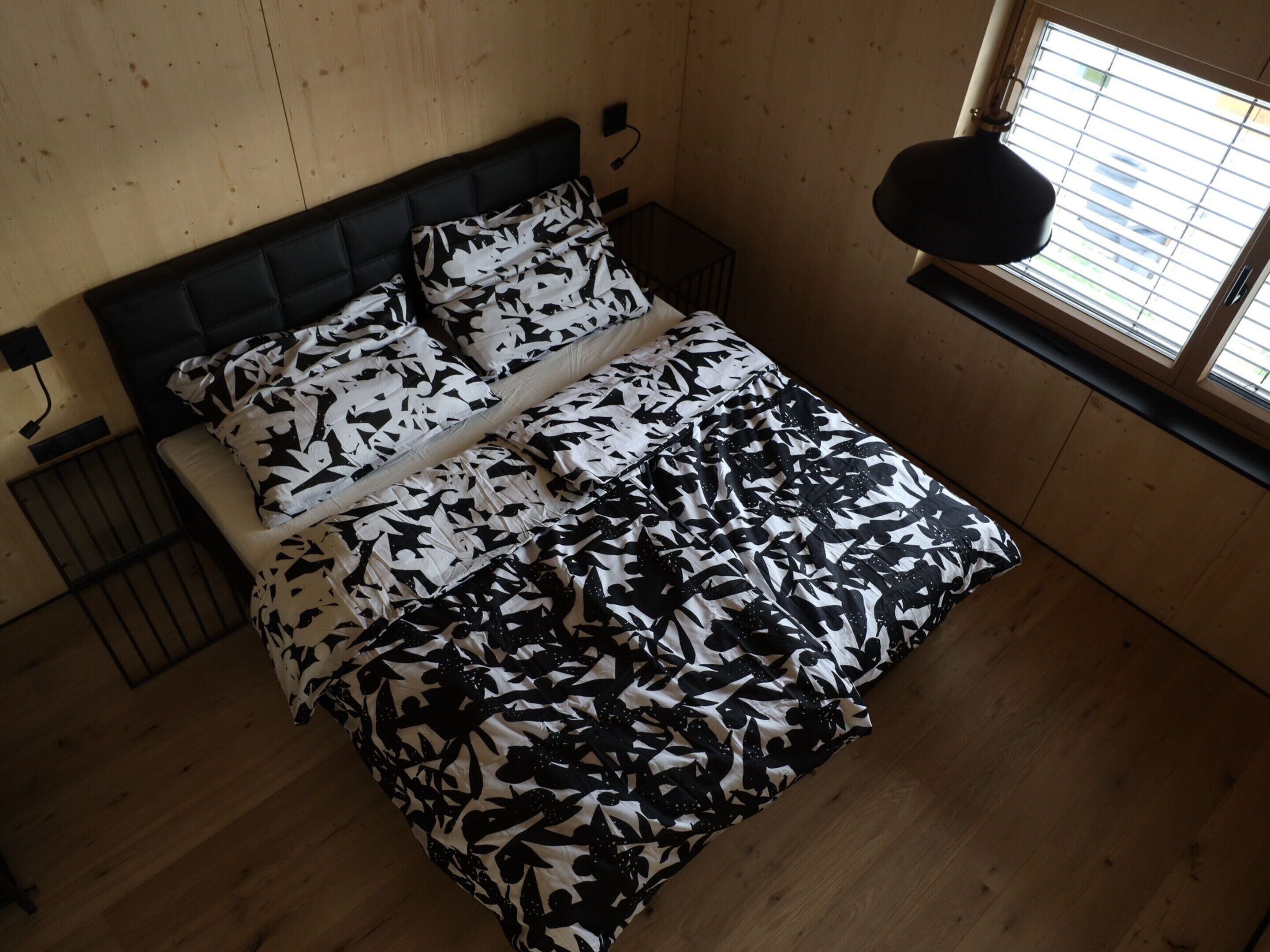 A bed with a black and white floral bedspread, a wooden headboard, and a window with blinds.