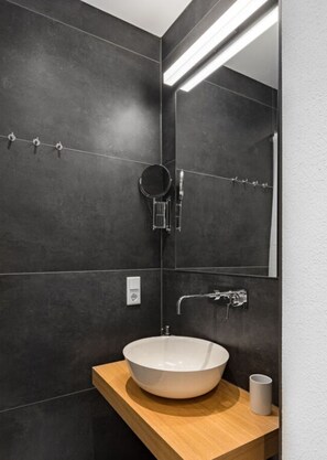 A modern bathroom with dark tiles, a white sink, a wooden countertop, a round mirror, and a towel rack. - in het Blauwe Huis - BG (Garmisch-Partenkirchen)