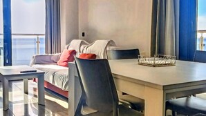 Une chambre d’hôtel moderne avec un lit, une table à manger et des chaises. Il y a une fenêtre avec des rideaux, ainsi qu’une petite table sur laquelle se trouve un récipient en verre.