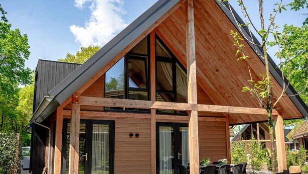 Una casa moderna de madera con techo inclinado, ventanas amplias y un patio con mobiliario exterior.
