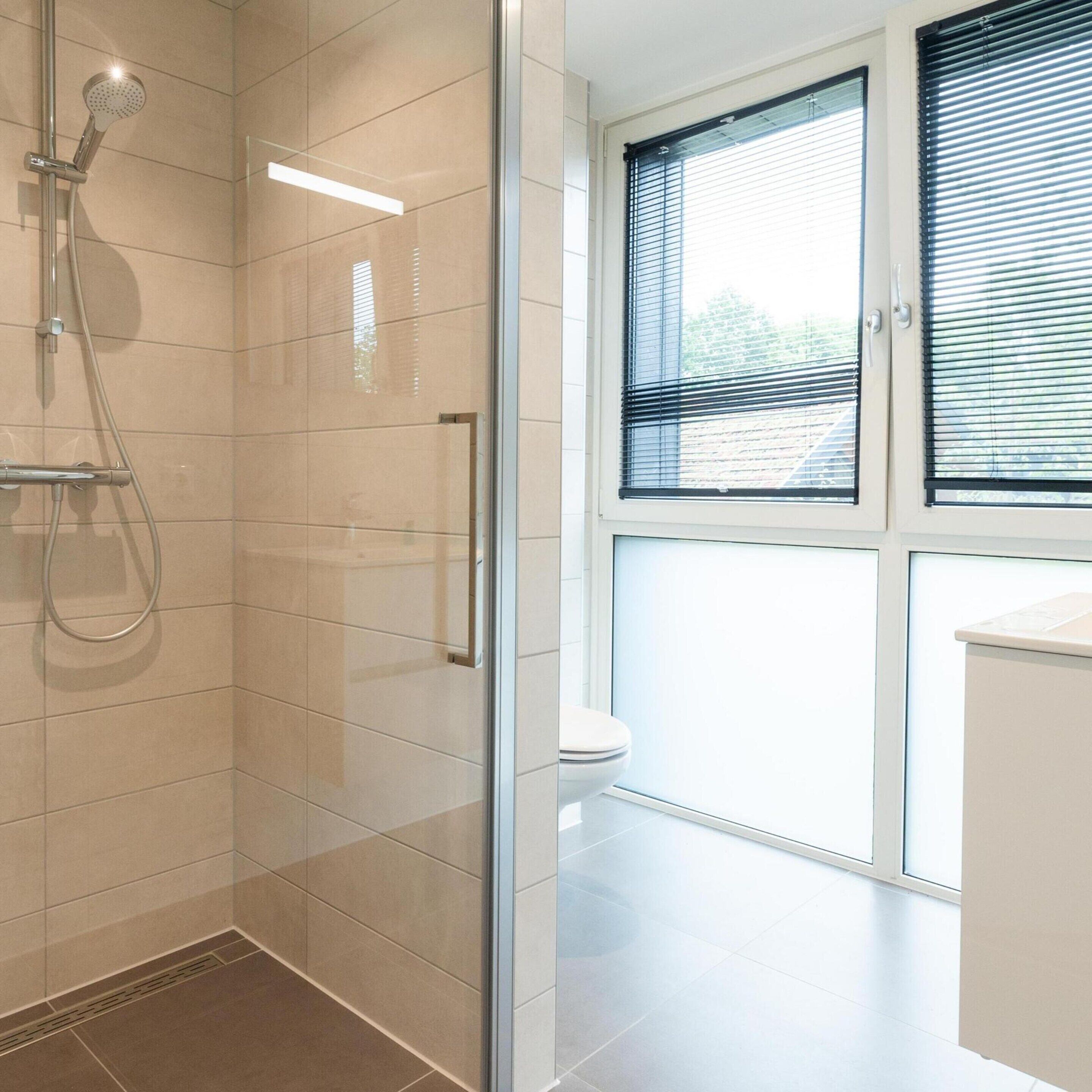 Un baño moderno con cabina de ducha de vidrio, inodoro y ventana con persianas.