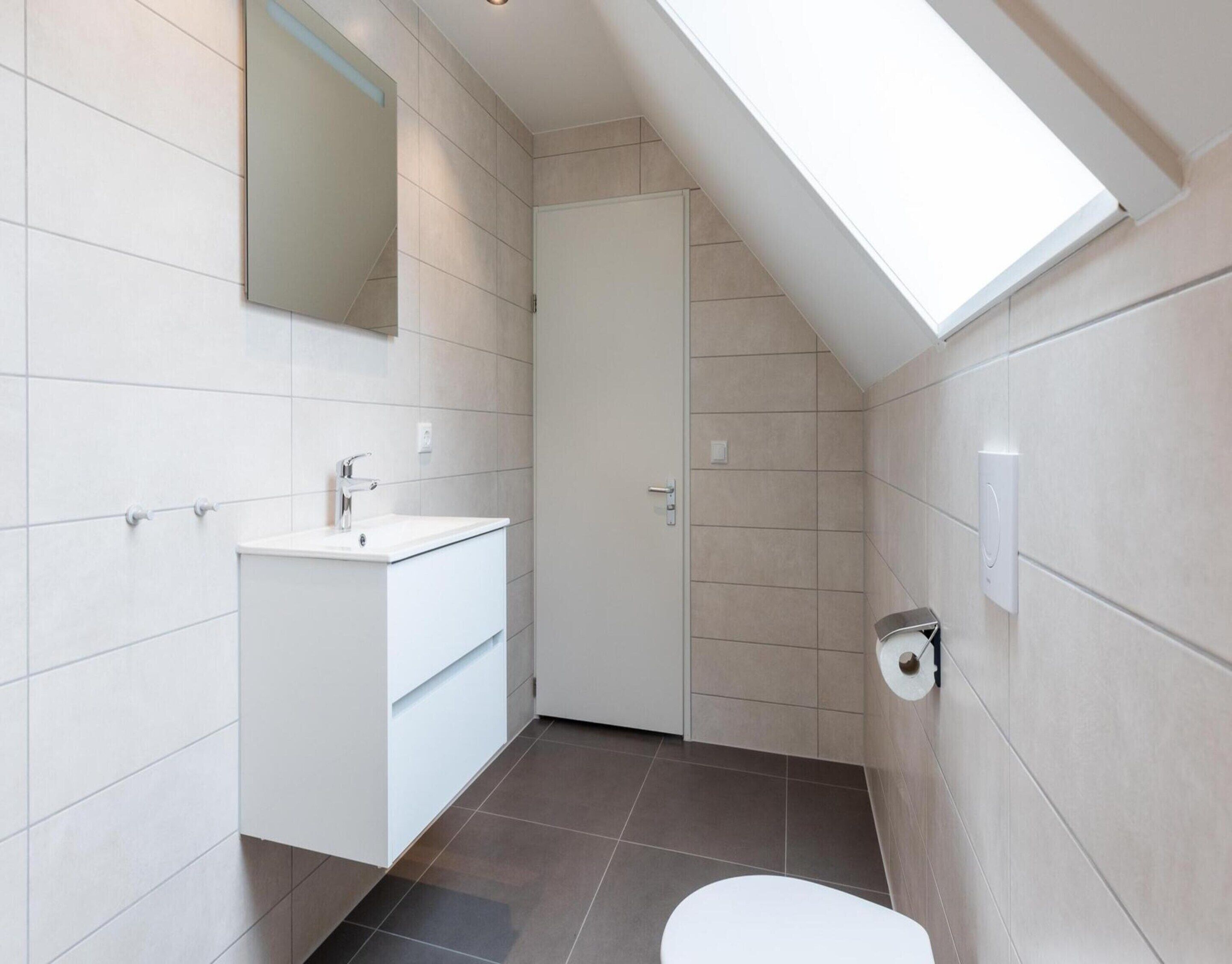 Un baño moderno con tragaluz, azulejos blancos, lavabo blanco y retrete.