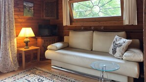 A cozy cabin interior with a sofa, a glass table, a lamp, a TV, and a window with a view of trees. - Blockhaus Mitten in der Natur (Eschede)