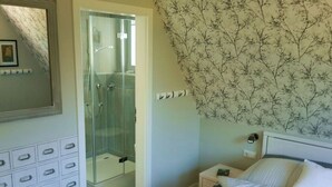 A bedroom with a bed, a nightstand, a dresser with drawers, a shower area, and a wall with a floral pattern. - Tonnihuus Comfortable Holiday Residence (Toenning)