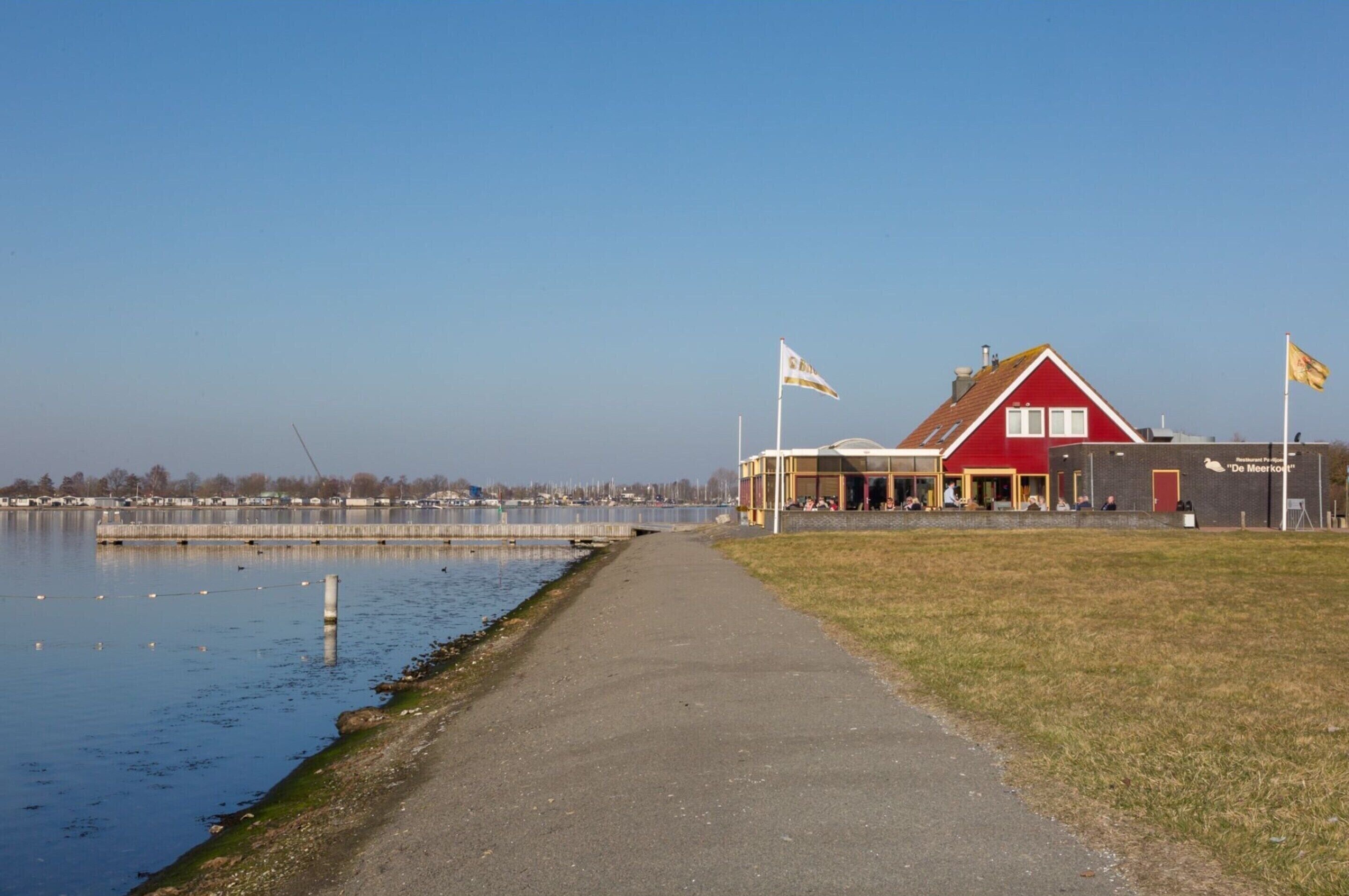A red building with a white sign, a pathway beside a body of water, and flags on poles.