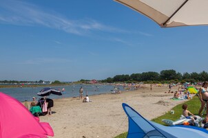 Una playa con gente relajándose y tomando el sol, tiendas de campaña, y una masa de agua al fondo.