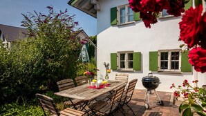 An outdoor patio with a wooden table and chairs, a barbecue grill, and a white building with green shutters. - Fisherman's House Comfortable Holiday Residence (Dießen am Ammersee)