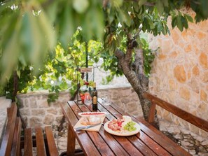 Une table en bois placée à l’extérieur, avec une bouteille de vin, un verre et une assiette de nourriture.