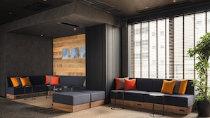A modern lounge area with dark sofas, a small round table, and a large window with blinds. - Nest Hotel Tokyo Yaesu (Tokyo)