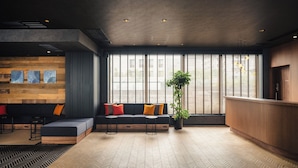 A modern lobby with a wooden reception desk, a black sofa with colorful cushions, a potted plant, and a herringbone-patterned floor. - Nest Hotel Tokyo Yaesu (Tokyo)
