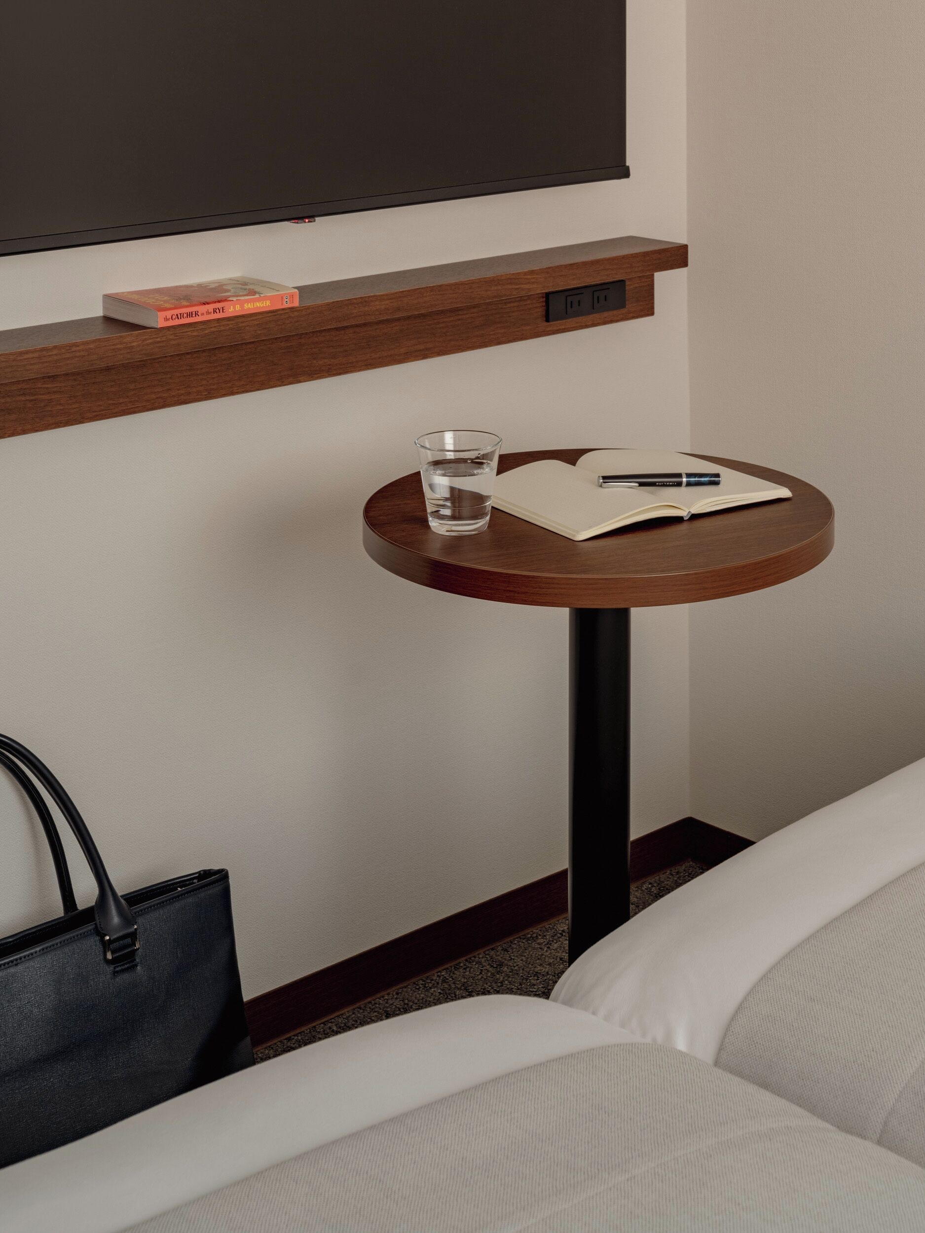 Habitación de hotel con una mesa redonda de madera, un vaso de agua, un cuaderno, un bolígrafo y un bolso negro.