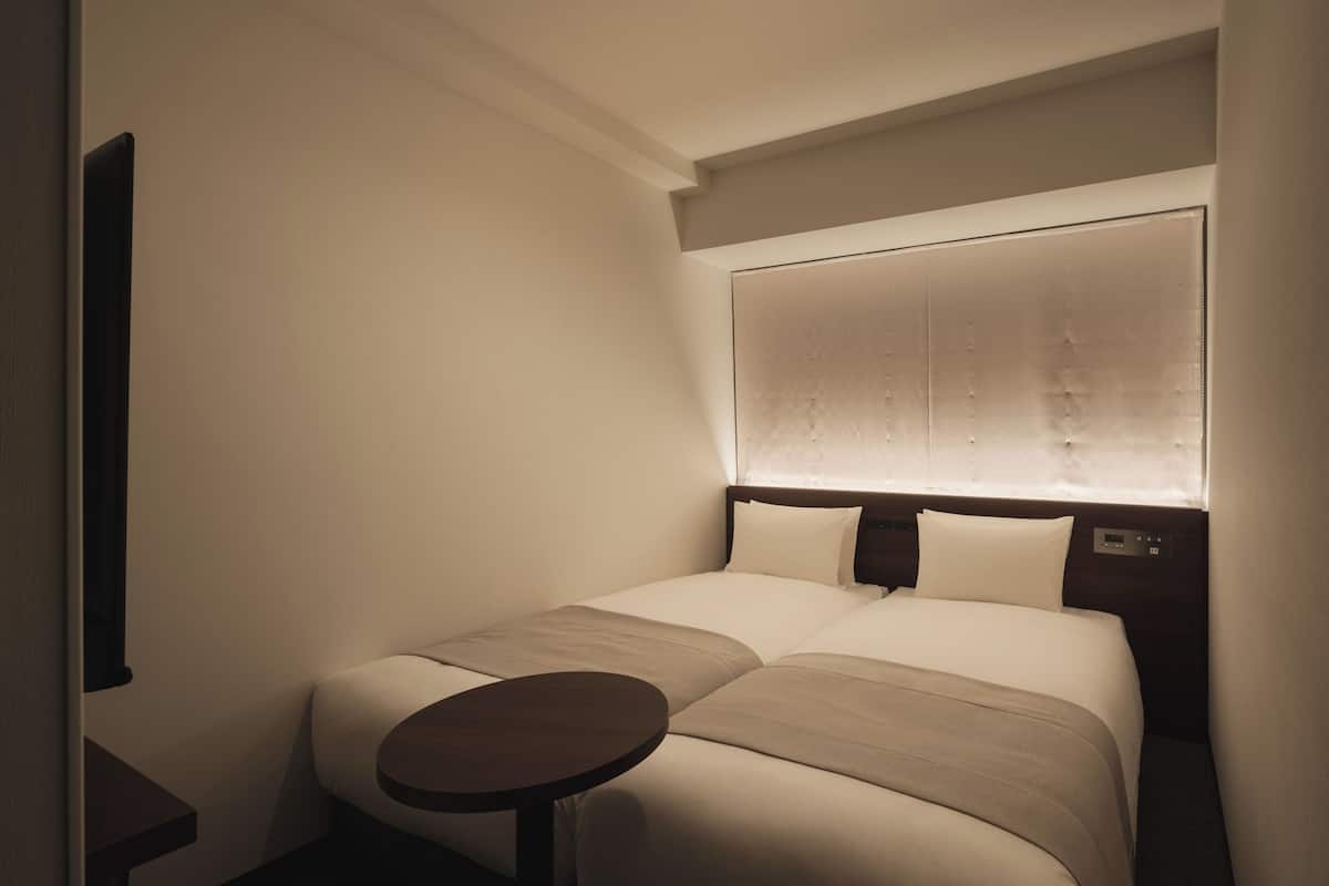 Una habitación de hotel con cama, mesita de noche y ventana con persianas.
