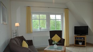 Un salon comprenant un canapé, un fauteuil et une table basse. On y trouve une fenêtre avec des rideaux, un radiateur et un téléviseur posé sur un meuble.