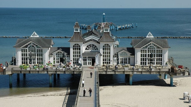 Une jetée sur laquelle se trouve un bâtiment blanc doté de grandes fenêtres et d’une entrée centrale, qui s’étend jusqu’à une plage de sable.