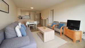 A living room with a grey sofa, a wooden coffee table, a TV stand, and a kitchen in the background. - Apartment Oostduinkerke Near the Beach (Koksijde)
