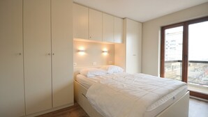 A bedroom with a bed, built-in wardrobe, and a large window with a city view. - Apartment Oostduinkerke Near the Beach (Koksijde)