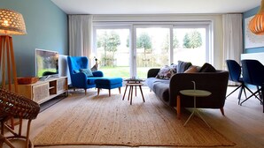 Uma sala de estar com uma poltrona azul, um sofá cinza, uma mesa redonda pequena e uma televisão em um armário de madeira.
