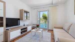 A modern living room with a flat-screen TV, a wooden cabinet, a white sofa, and a balcony with a view of greenery and water. - Apartments Ema-two Bedroom Apartment With Sea View (Ploce)