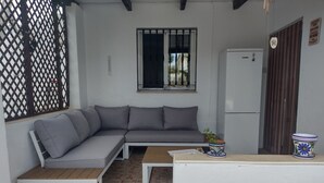 A compact living space with a grey sectional sofa, a small wooden table, a white wall, a window with black bars, a white refrigerator, and a decorative pot.