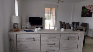 A modern living room with a central island, a flat-screen TV, and a dining area with chairs.