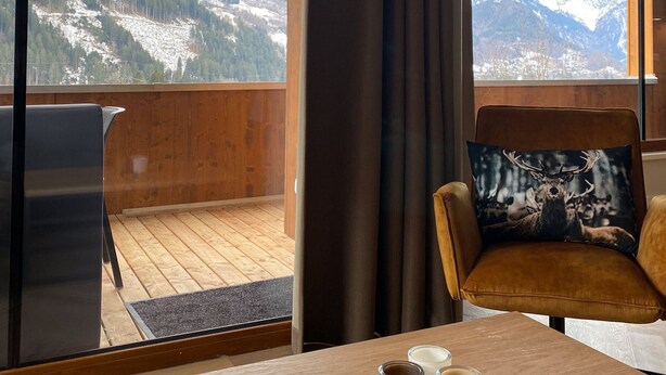Una habitación con piso de madera, una mesa con dos tazas, una silla con un cojín de ciervo y vistas a montañas nevadas a través de amplias ventanas.