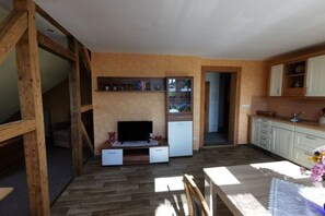 A compact living space with a wooden floor, a TV on a built-in shelf, a small kitchen area, and a dining table with a vase of flowers. - Holiday Apartment Mountain Idyll (Jonsdorf)