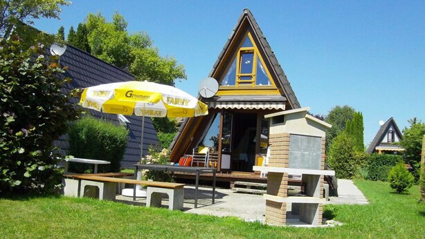 Een klein, gebouwtje met een puntig dak, een terras, zitplaatsen buiten en een grote gele parasol.