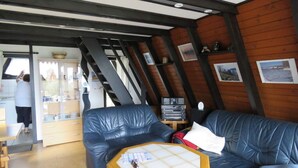 Een gezellige kamer met een houten plafond, een zwarte leren bank, een houten salontafel en een keukenhoek waar iemand kookt.