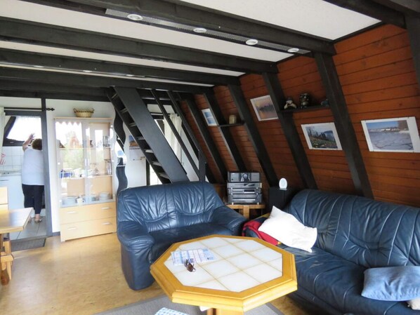 A cozy room with a wooden ceiling, a black leather sofa, a wooden coffee table, and a kitchen area with a person cooking.