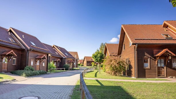A row of wooden cabins with pitched roofs, a paved walkway, and a grassy area with a wooden fence. - Holiday Home in Hasselfelde (Oberharz am Brocken)