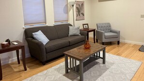 A living room with a grey sofa, a grey armchair, a wooden coffee table, and a patterned rug. There are two windows with blinds, a wall-mounted air conditioner, and a framed botanical print on the wall.