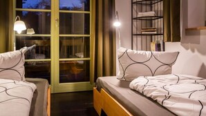 Et soveværelse med to senge, et vindue med gardiner, en sengebordlampe og en metalkasse med bøger.